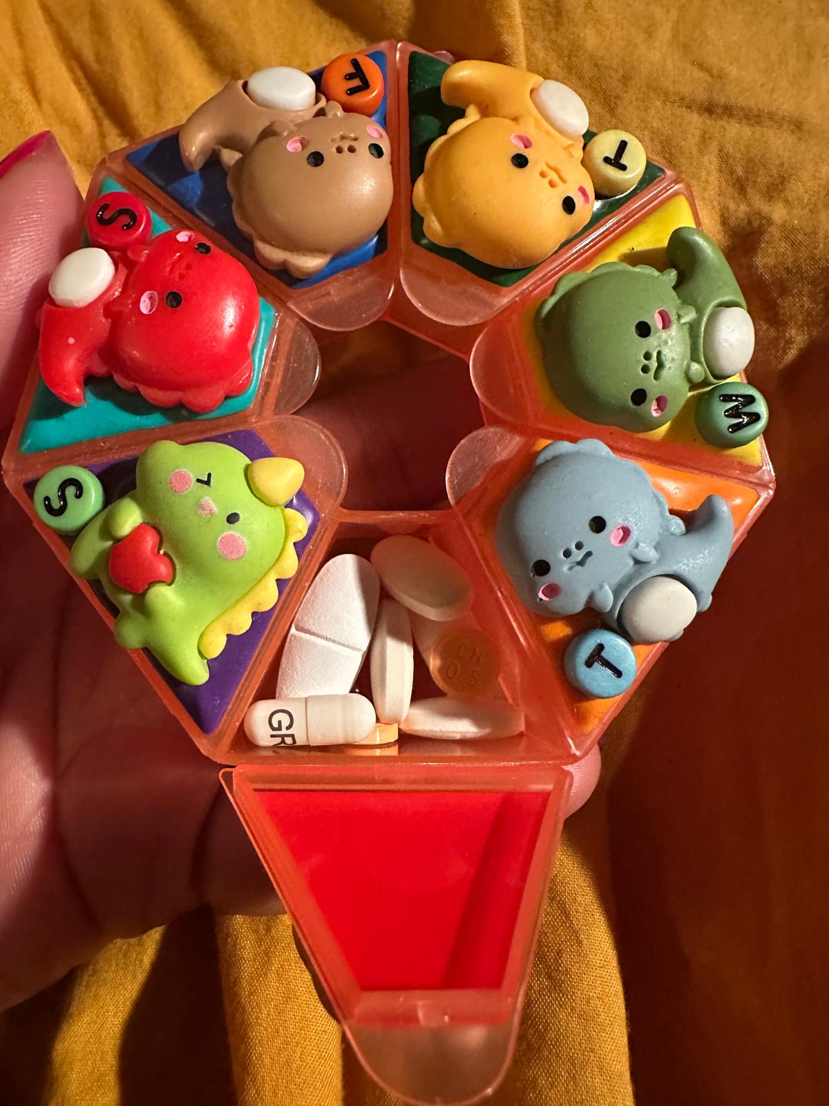 A pill dispenser with lids that have baby dinosaurs on them. One is open to show a multitude of pills.